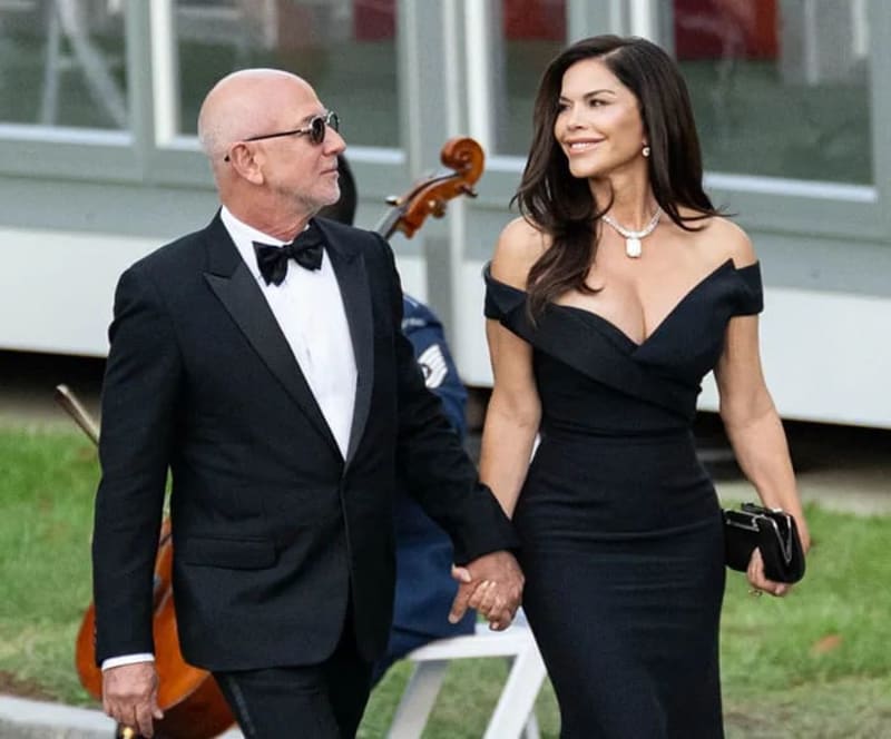 Two elegantly dressed people hold hands while walking outdoors at a formal event, with a violin visible in the background.