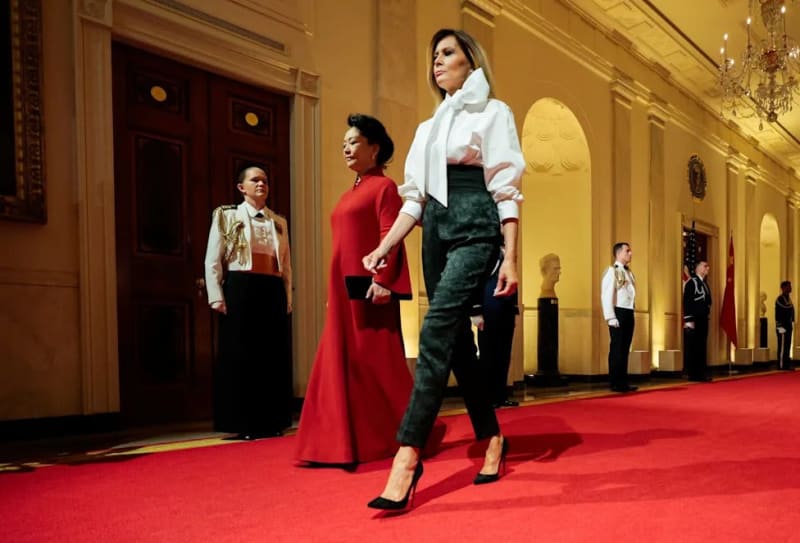 Two women walking down red-carpeted White House hallway, one in white blouse with dark trousers, one in red gown.
