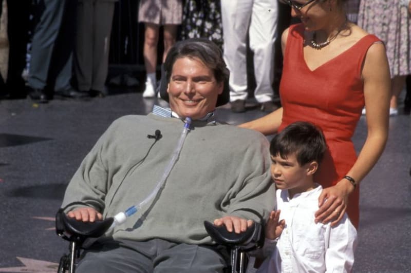 A man in a wheelchair wearing a gray jacket smiles while posing with a woman in a red dress and young boy at an outdoor event.
