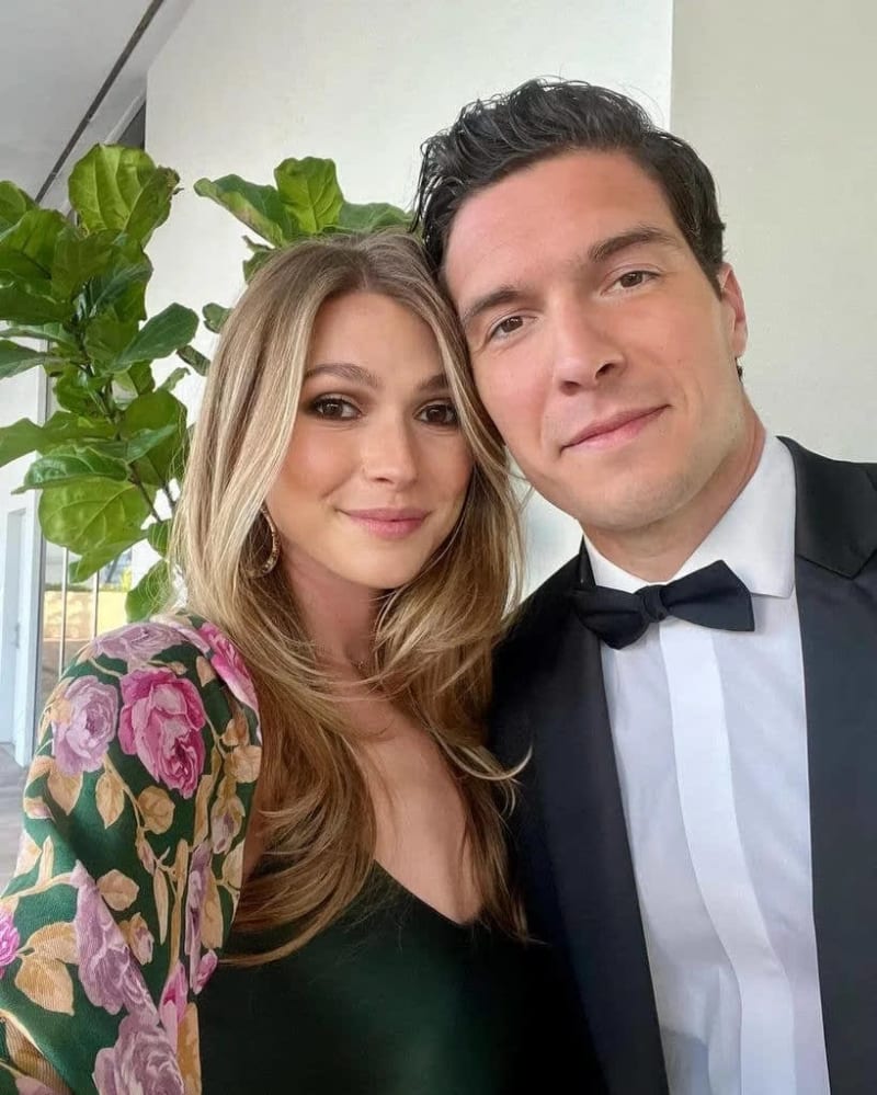 Two people posing together for a photo, with one wearing formal black tie attire and the other in a floral patterned top.