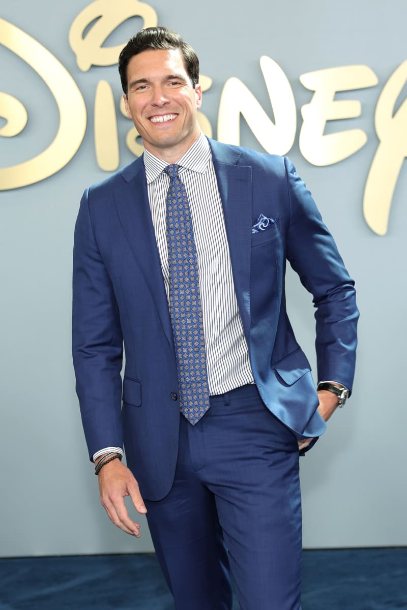 A man in a navy blue suit and patterned tie smiles at a Disney event premiere.