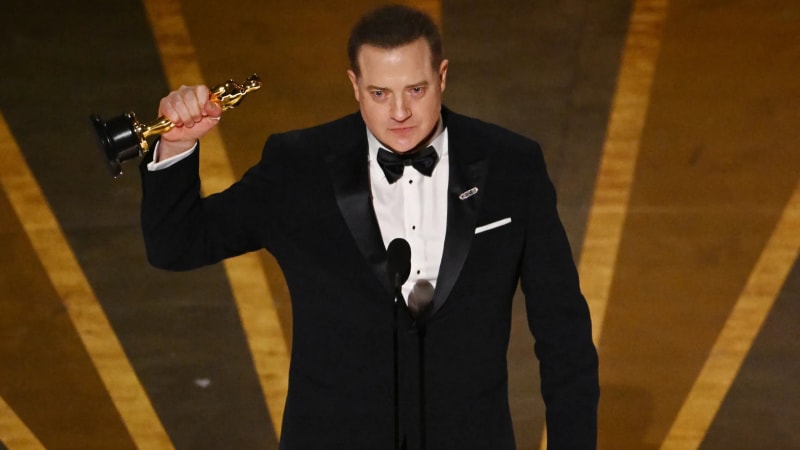 Man in black tuxedo holding Oscar statuette aloft at podium on stage.