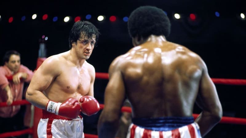 Two male boxers in red gloves and trunks facing off in a boxing ring.