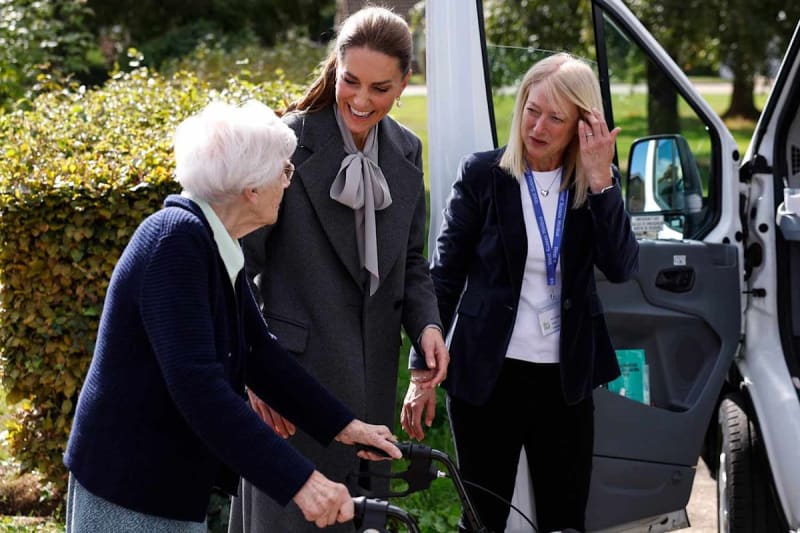 Three women assist an elderly woman into a white van in a garden setting on a sunny day.