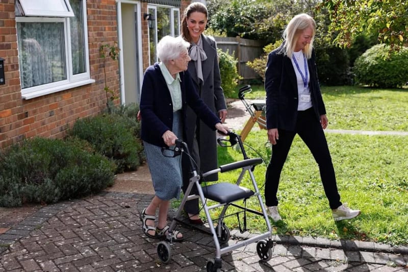 An elderly woman using a walker is assisted by two younger women in business attire while walking along a brick pathway.