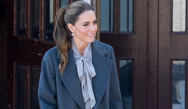 A woman in a charcoal blazer and gray bow tie smiles while standing against a dark wooden building facade.