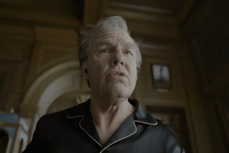 An older man with gray hair wearing a dark jacket looks upward in what appears to be an ornate interior space.