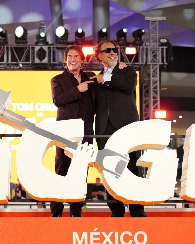 Two men in dark suits smiling on a stage above a large film logo and México signage.