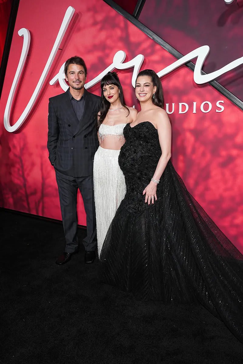 Man in dark suit, woman in white sequin gown, woman in black strapless gown, red carpet backdrop.