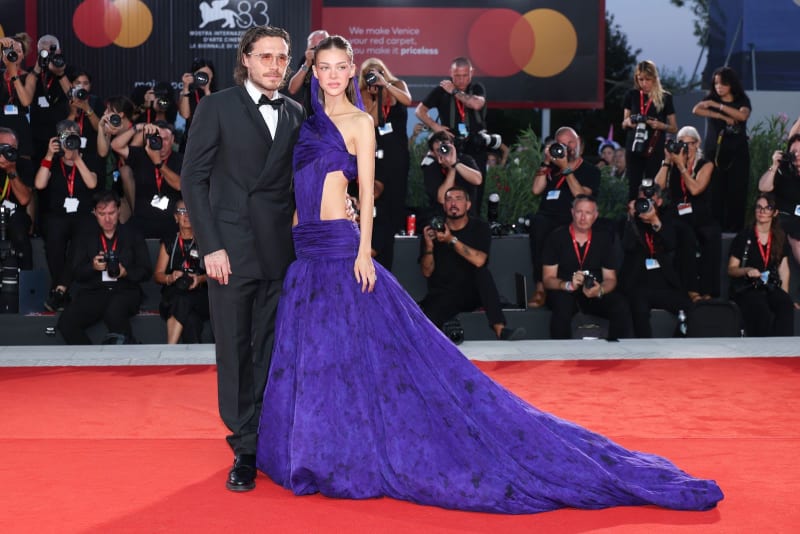 Two people on a red carpet at a film festival; man in black tuxedo and woman in purple gown with train, surrounded by photographers.