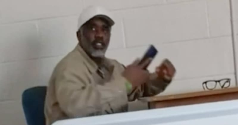 A man wearing a white cap and tan jacket sits at a desk while holding a marker, appearing to teach or present.