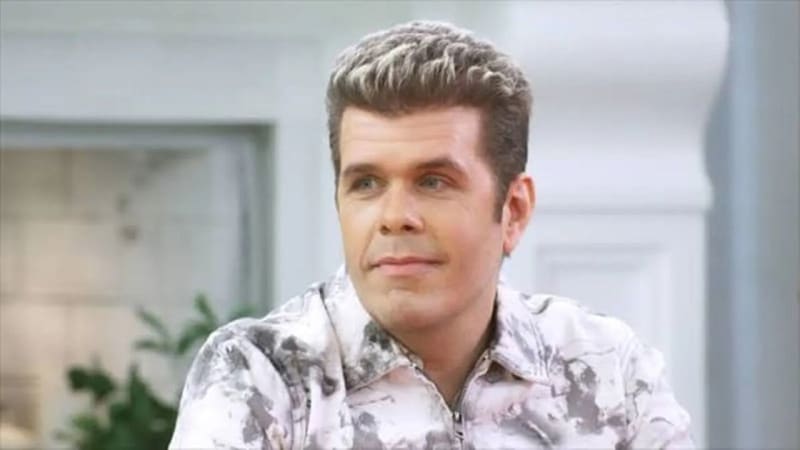 A man with styled blonde hair wearing a patterned white and gray shirt in an indoor setting with plants visible in the background.