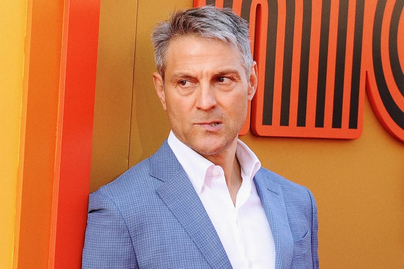 Man in blue blazer and white shirt, standing at orange and gold branded backdrop.