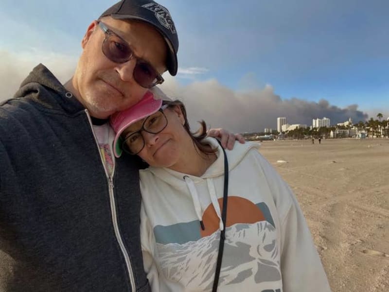 Two people posing together on a beach with coastal buildings and smoke visible in the background.