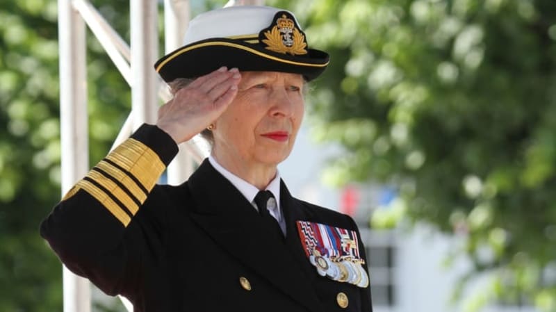 A high-ranking naval officer in dress uniform salutes while wearing a captain's hat with gold insignia and sleeve stripes.