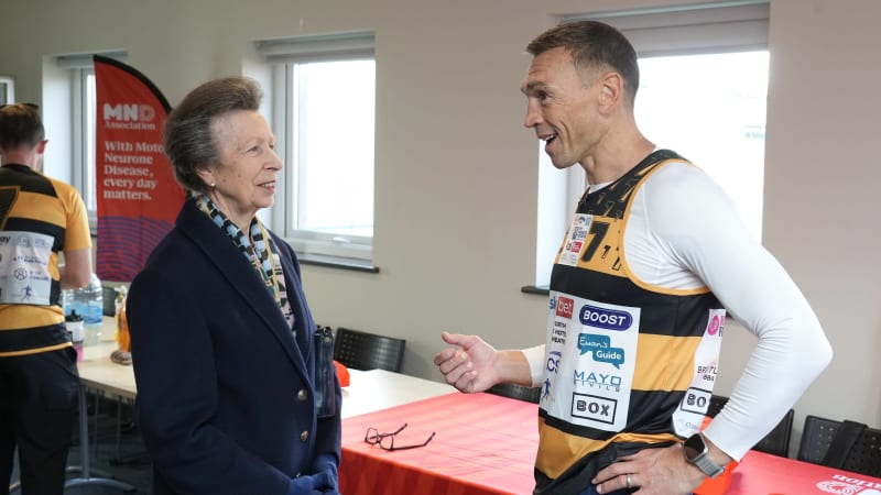 A woman in formal navy blazer and medal converses with a smiling man wearing a yellow and black sports jersey with sponsor logos.