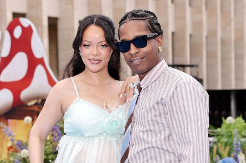 Two people posing together at an outdoor event, with one wearing a light blue and white dress and the other in a striped shirt and sunglasses.