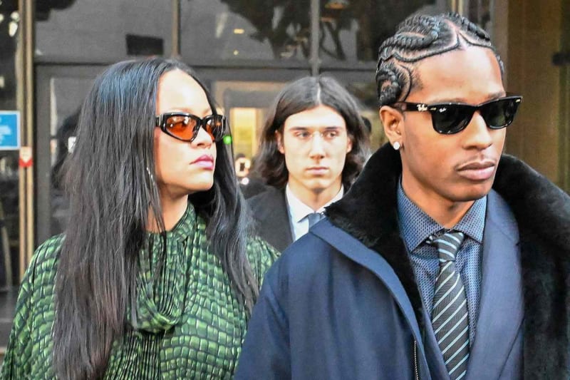 Woman in green patterned top and tinted sunglasses walks beside man in navy coat and tie, city street.