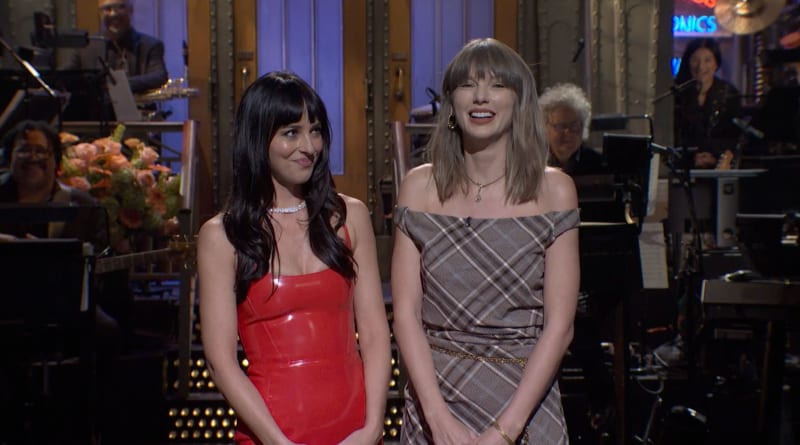 Two women stand on a stage in elegant dresses, smiling at each other during what appears to be a television performance or appearance.