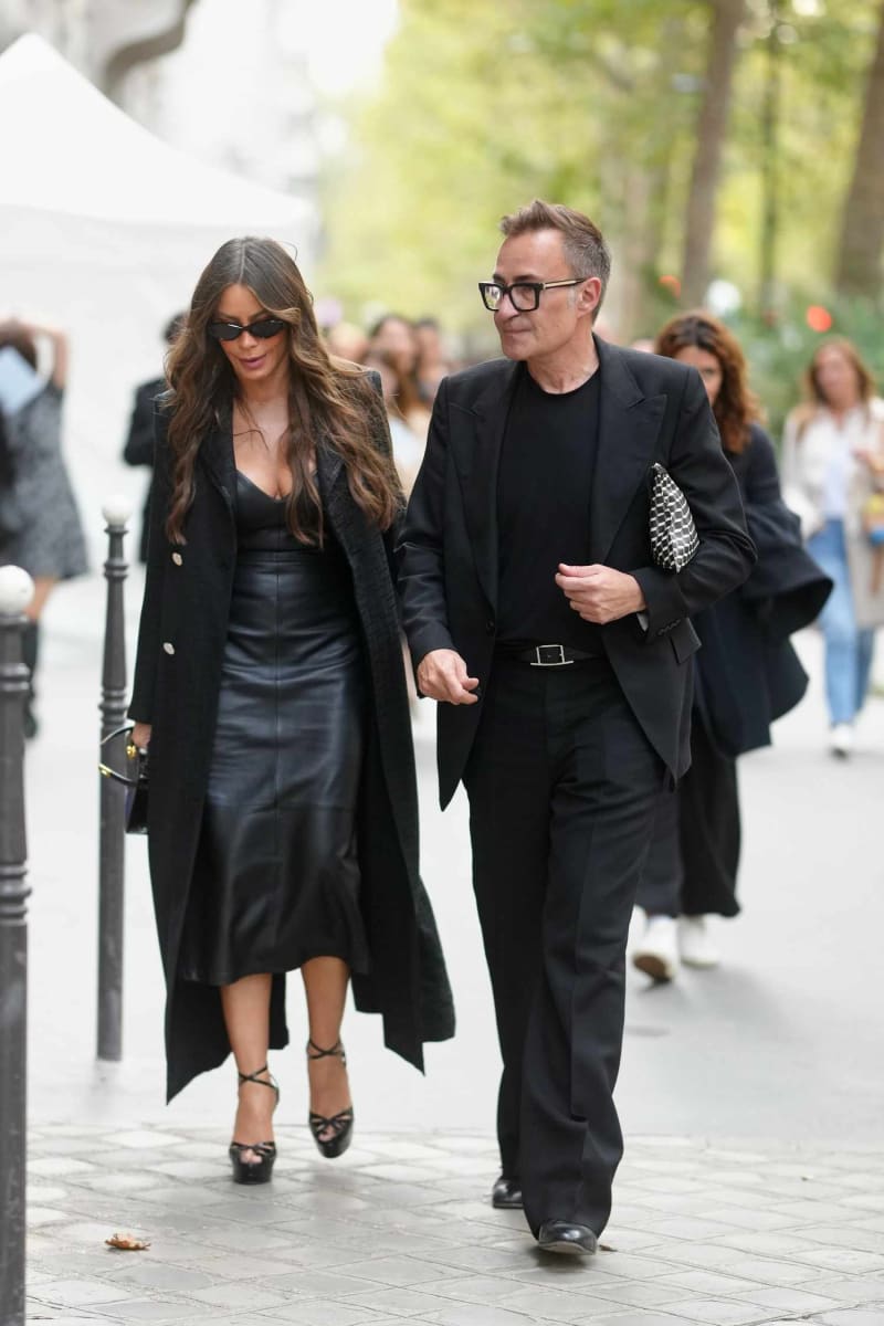 Woman in black leather skirt and long coat walking with man in black suit on city street.