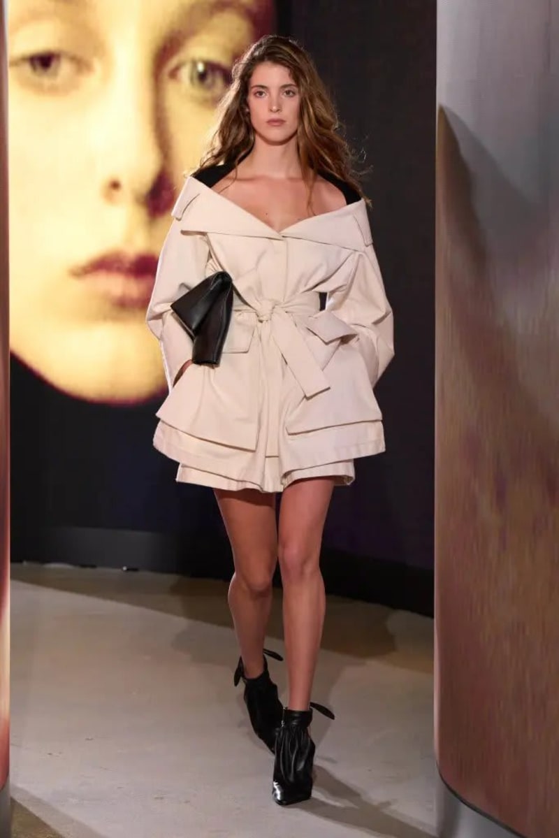 Woman in cream off-shoulder belted jacket and black boots, walking runway.