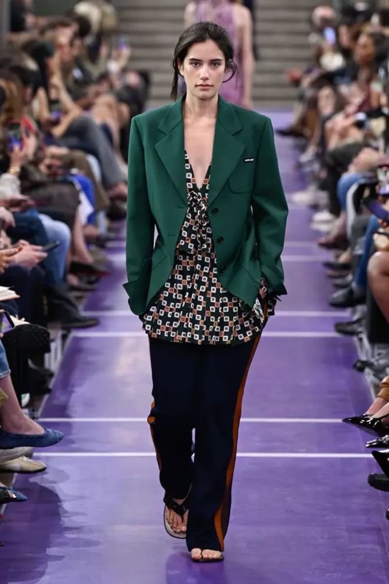 A model walks down a fashion runway wearing a green blazer over a patterned blouse and black pants with orange side stripes.