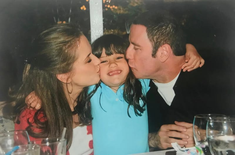 Woman in red floral top and man in black jacket both kissing cheeks of smiling child in blue polo, seated at dining table.