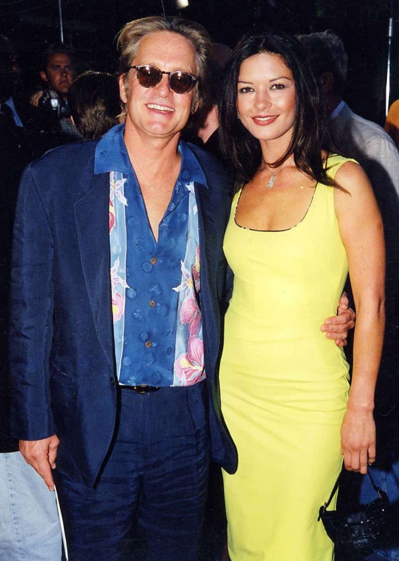 Man in blue blazer and floral shirt with sunglasses standing with woman in yellow dress at an event.