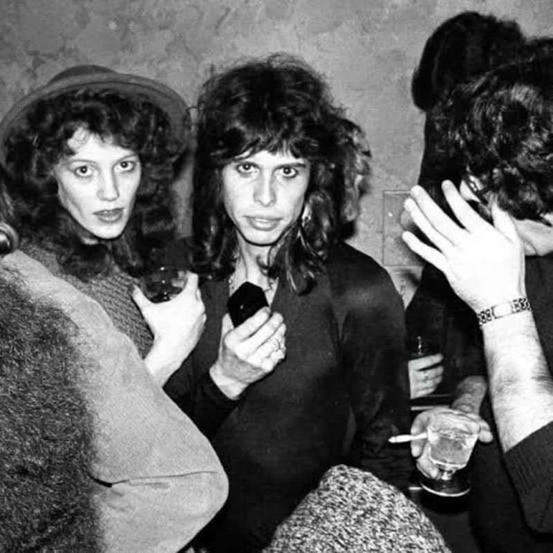 Woman in hat and man with long hair holding drink at indoor party, black and white photo.