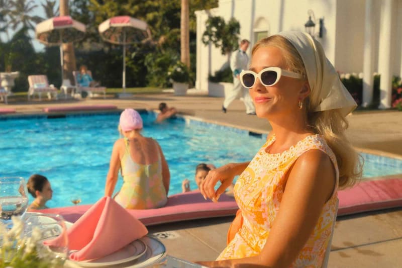 Woman in yellow patterned dress, white cat-eye sunglasses and head scarf, seated poolside at a retro estate.