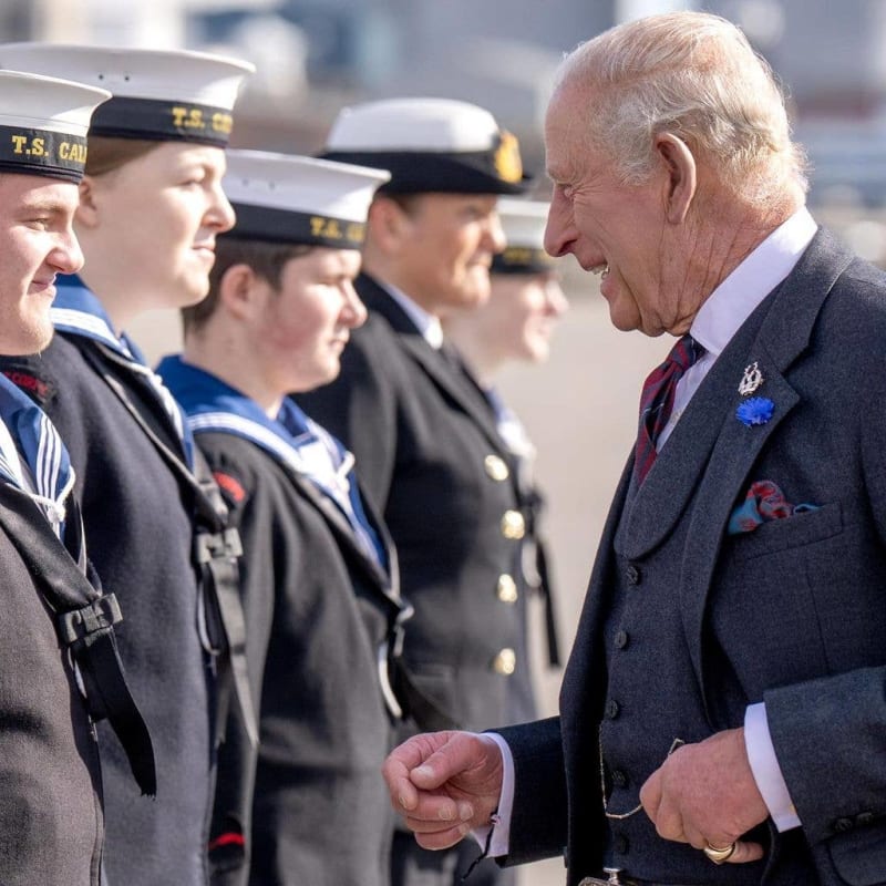 An elderly man in a formal suit speaks with military personnel wearing naval uniforms and caps marked "T.S. CAD."