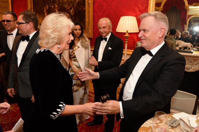 A woman in black velvet greets a man in a tuxedo at a formal palace reception with ornate red walls and gold décor.