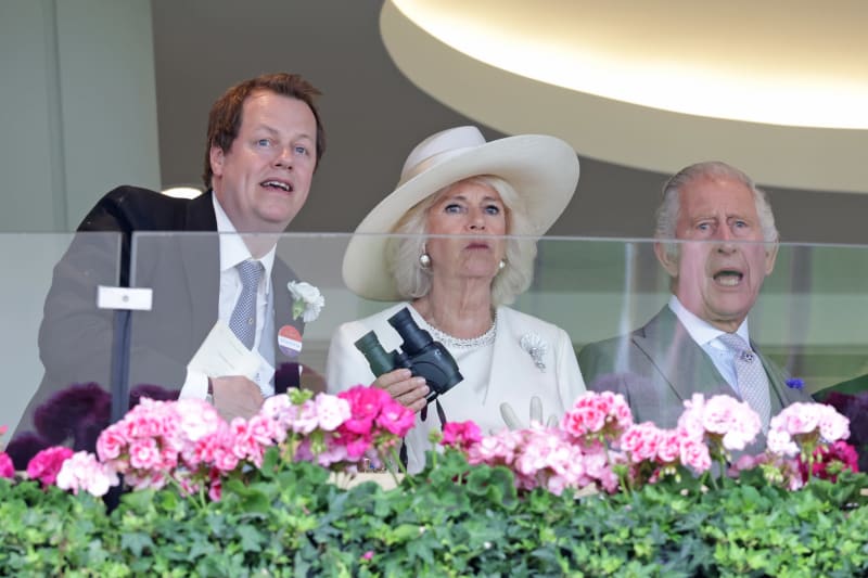 Three elegantly dressed people watch from a balcony decorated with pink and magenta flowers at what appears to be a formal event.
