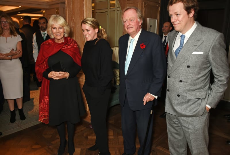 Formally dressed guests at an indoor event, including a woman in a red shawl and black dress and men in business suits.