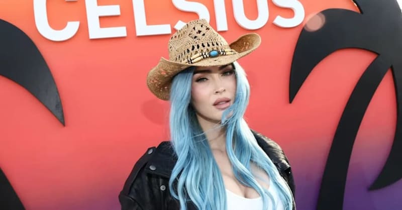 Woman with long light blue hair wearing a black leather jacket and straw cowboy hat at a red event backdrop.
