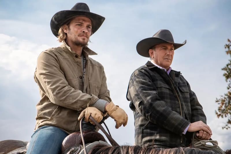 Two men in cowboy hats on horseback, one in tan jacket with gloves, one in plaid jacket, outdoors.