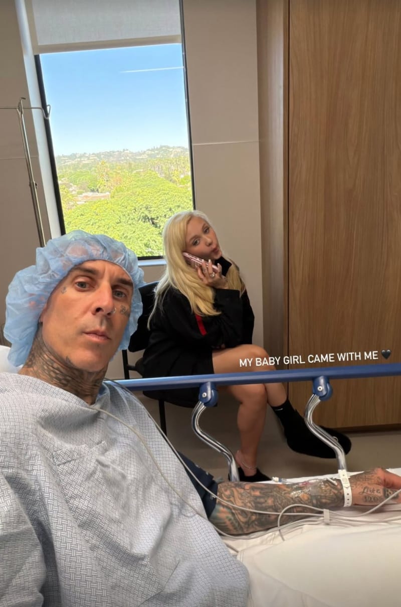 Tattooed man in surgical cap and hospital gown on bed; woman in black jacket on phone behind him, hospital room.
