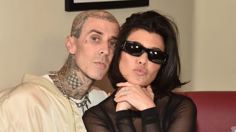 Two people posing together indoors; person on left has extensive facial tattoos and neck tattoos wearing a cream jacket, person on right wears black sunglasses and black clothing.