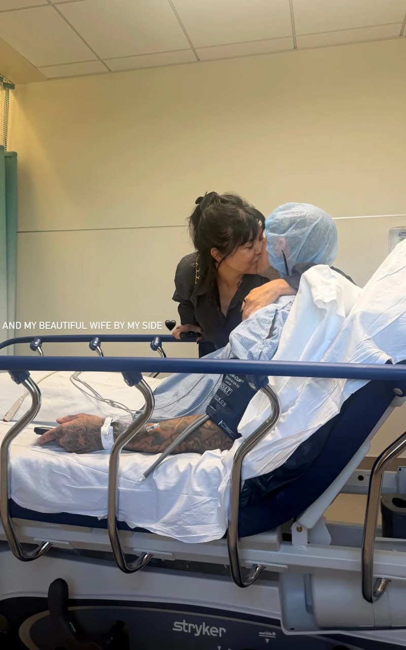 Woman in black top kissing tattooed man in surgical cap on hospital bed, hospital room.