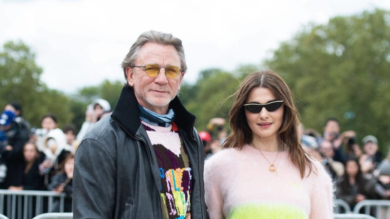 Two people stand together outdoors at a public event, with crowds and trees visible in the background.