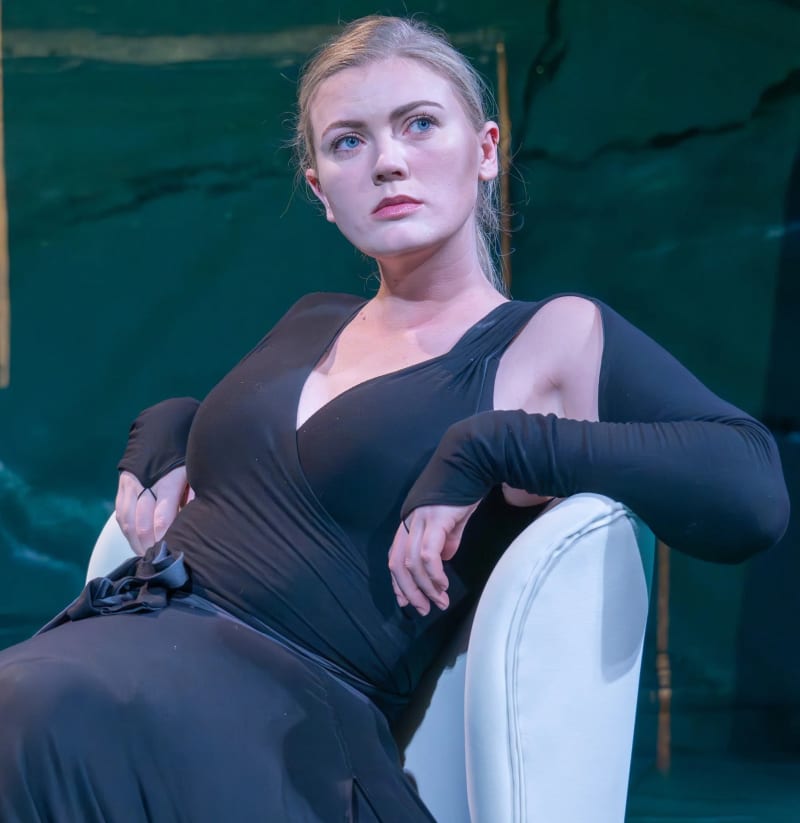 Woman in black long-sleeve cutout dress, seated in white chair against green backdrop.