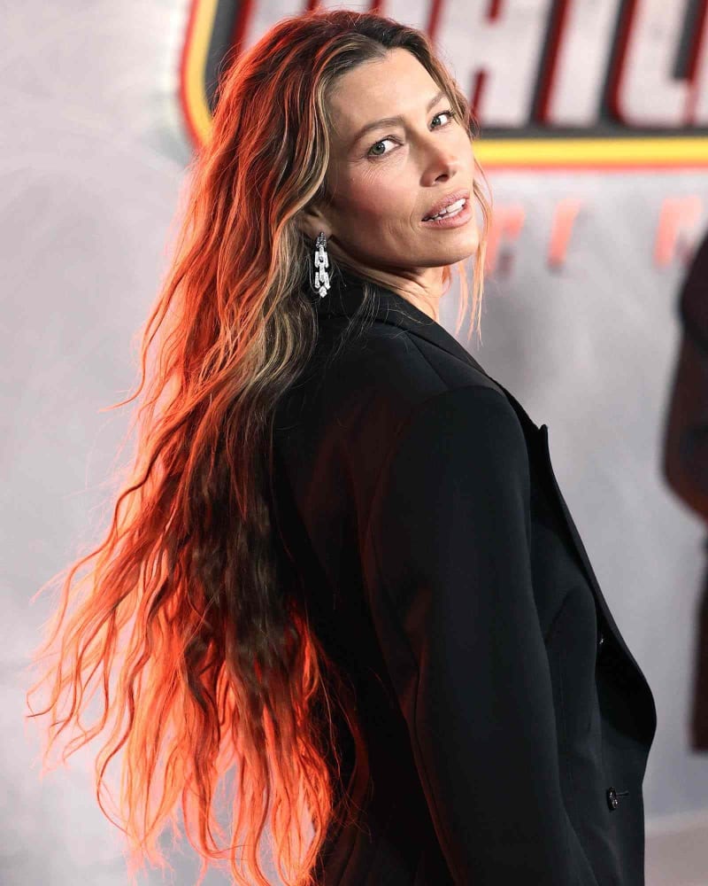 Woman in black blazer with long red-tinted hair, glancing over shoulder at premiere backdrop.