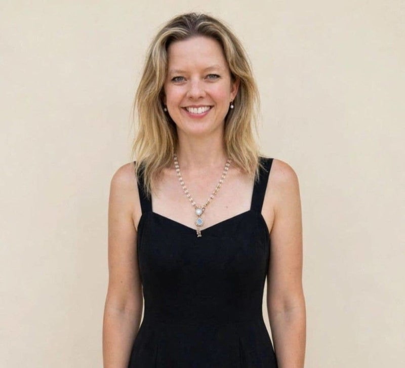 A woman with shoulder-length blonde hair smiling at the camera, wearing a black sleeveless dress and pearl necklace.