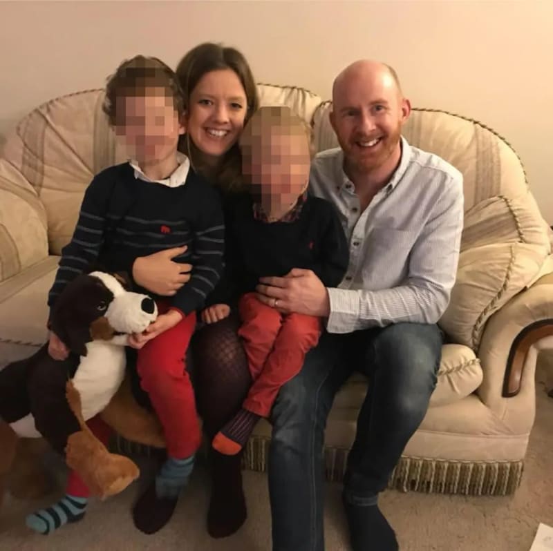 Woman and man seated on sofa holding two young children with pixelated faces, with a stuffed dog toy, indoors.