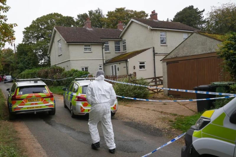 Forensic officer in white suit walking toward white cottage behind police cordon with two police cars, rural setting.