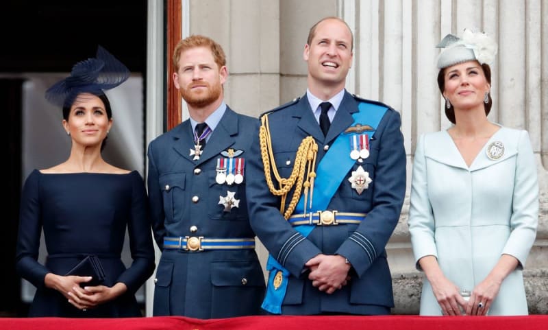 Four people stand on a balcony: a woman in black, two men in military dress uniforms, and a woman in white with a hat.