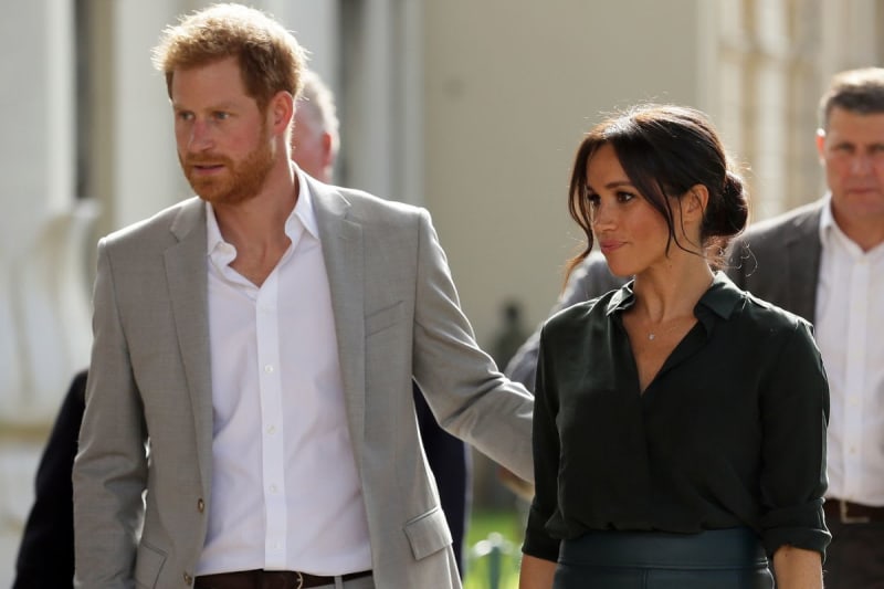A man in a beige blazer and white shirt walks alongside a woman in a black blouse in a formal hallway setting.