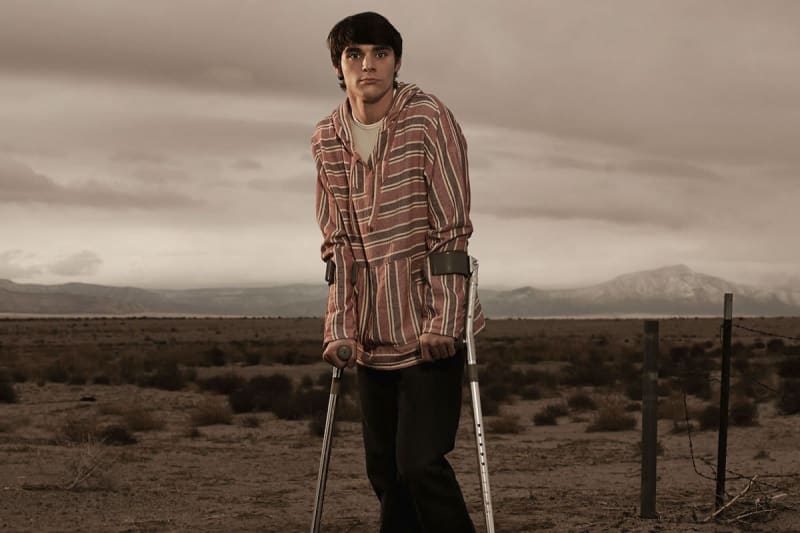 Young man in striped hoodie standing on crutches in desert landscape.