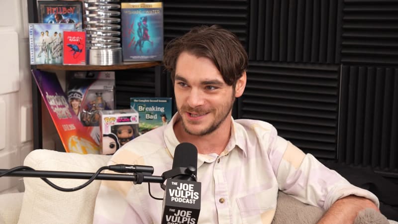 Man in striped white shirt seated at podcast microphone, studio shelving in background.