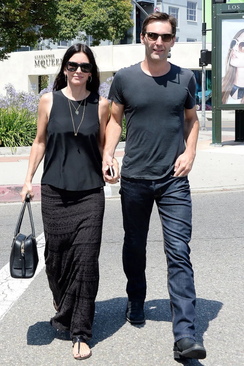 Woman in black top and maxi skirt holding hands with man in grey t-shirt and jeans, walking on sunny street.